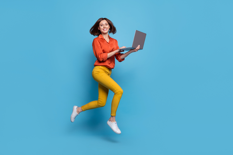 Woman jumping in the air holding a laptop against an aqua-colored background.