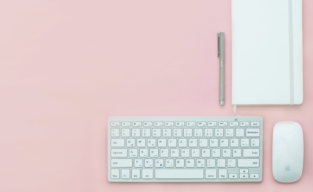 Keyboard, journal, pen, and mouse on pink desktop.